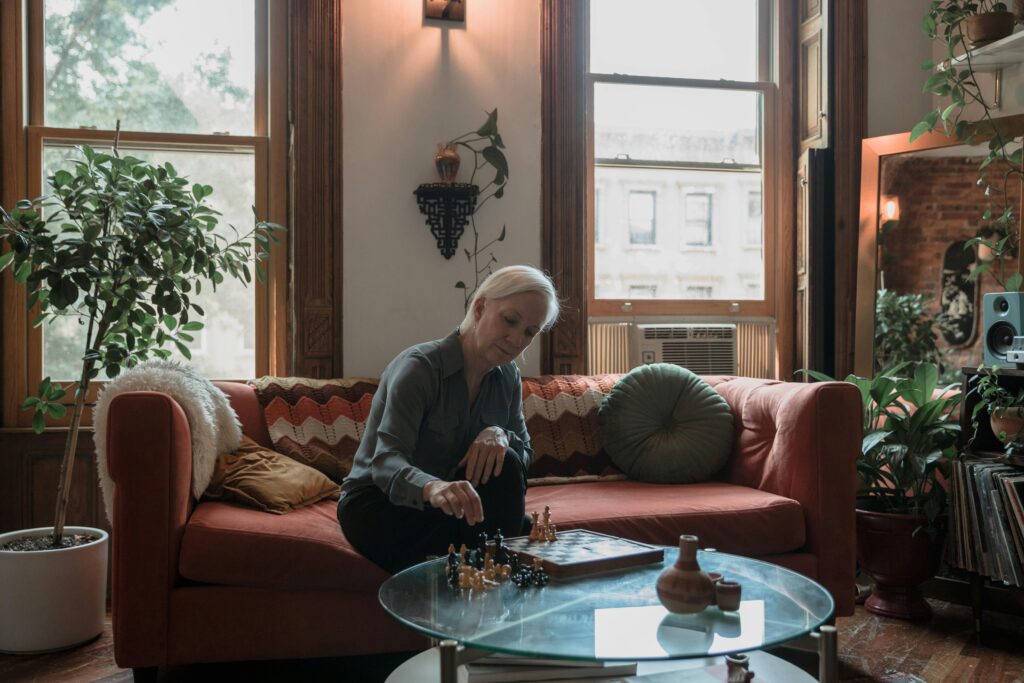 Elderly woman enjoying chess game on a sofa in a stylish, plant-filled room.
